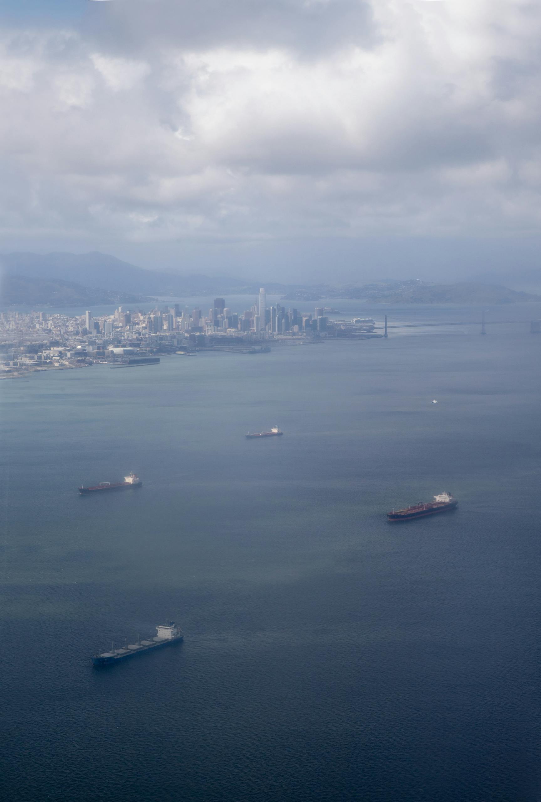 Aerial shot of cargo ships in San Francisco Bay with city skyline and iconic bridge in the background.
