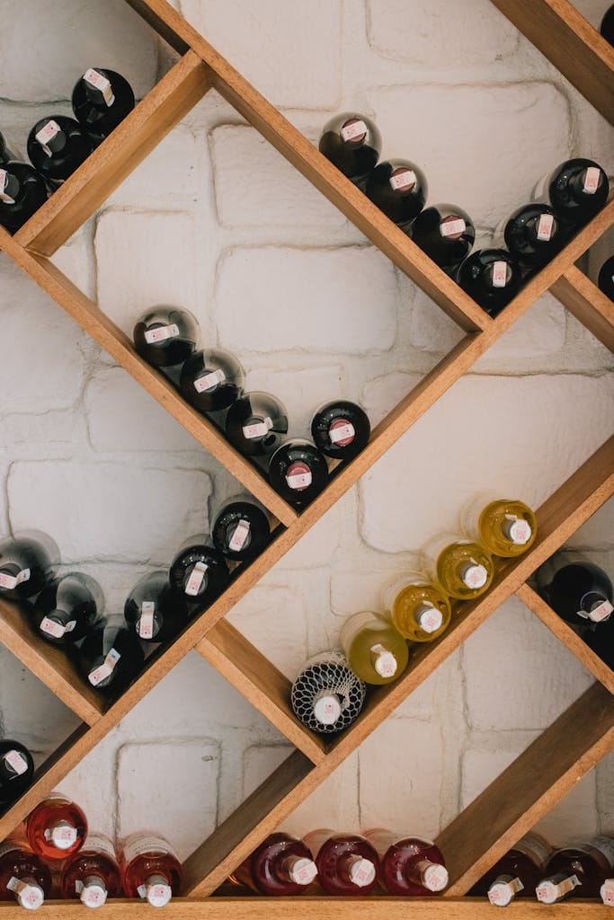 Rows of wine bottles on a wooden diagonal shelf against a brick wall.