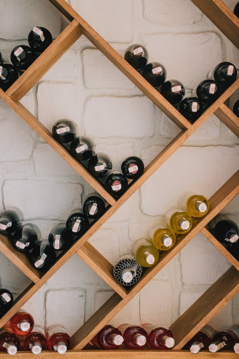 Rows of wine bottles on a wooden diagonal shelf against a brick wall.