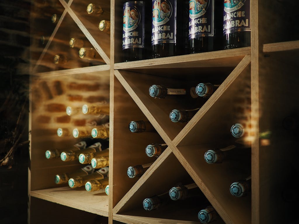 Wooden shelves in a wine cellar showcasing beer and wine bottles with a brick wall backdrop.