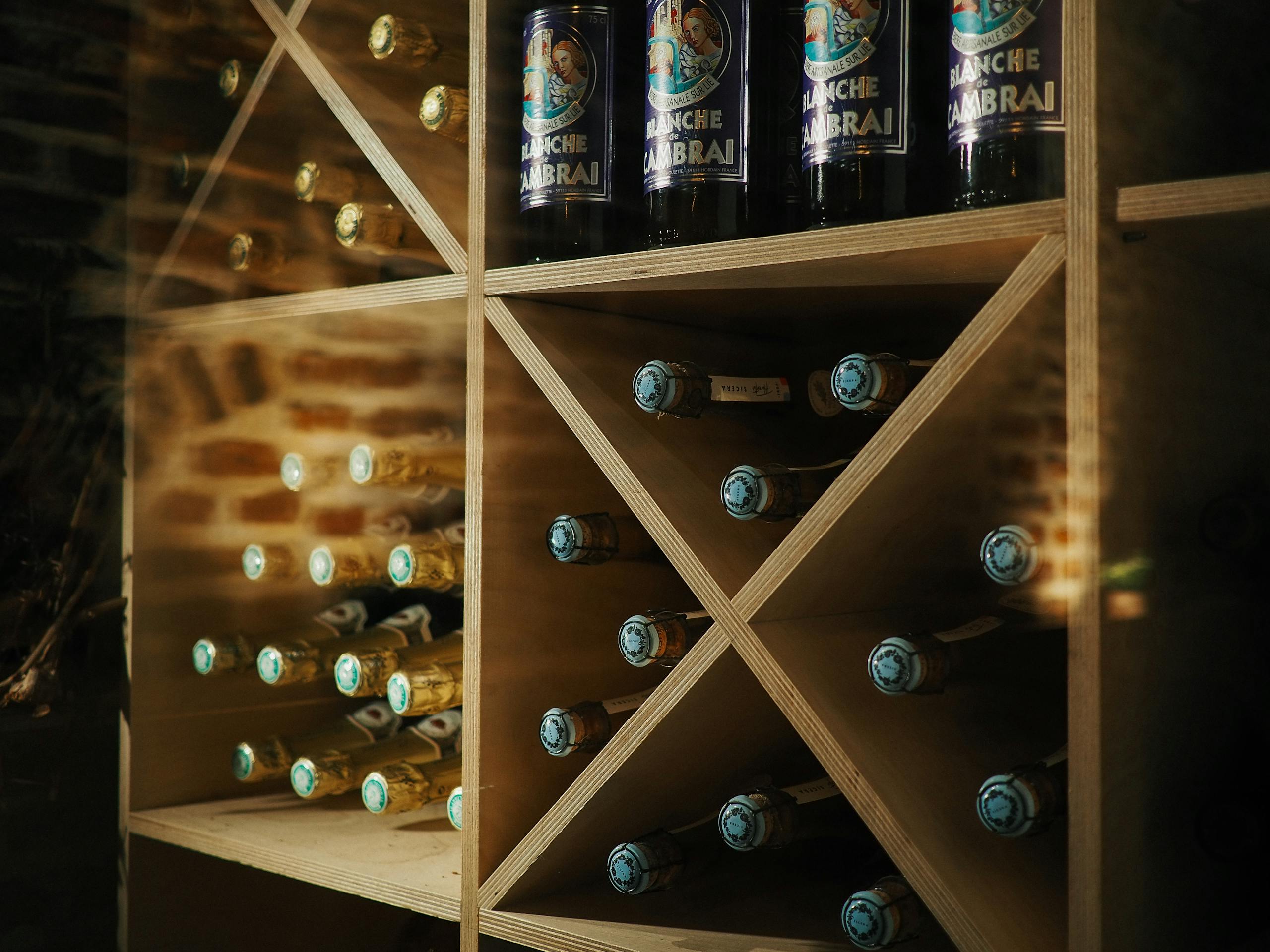 Wooden shelves in a wine cellar showcasing beer and wine bottles with a brick wall backdrop.